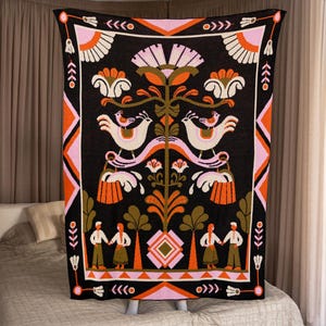 May include: A black blanket with a colorful folk art design. The blanket features white birds, orange and pink flowers, and figures holding hands. The border has geometric patterns in orange, pink, and black. The blanket is displayed on a bed.
