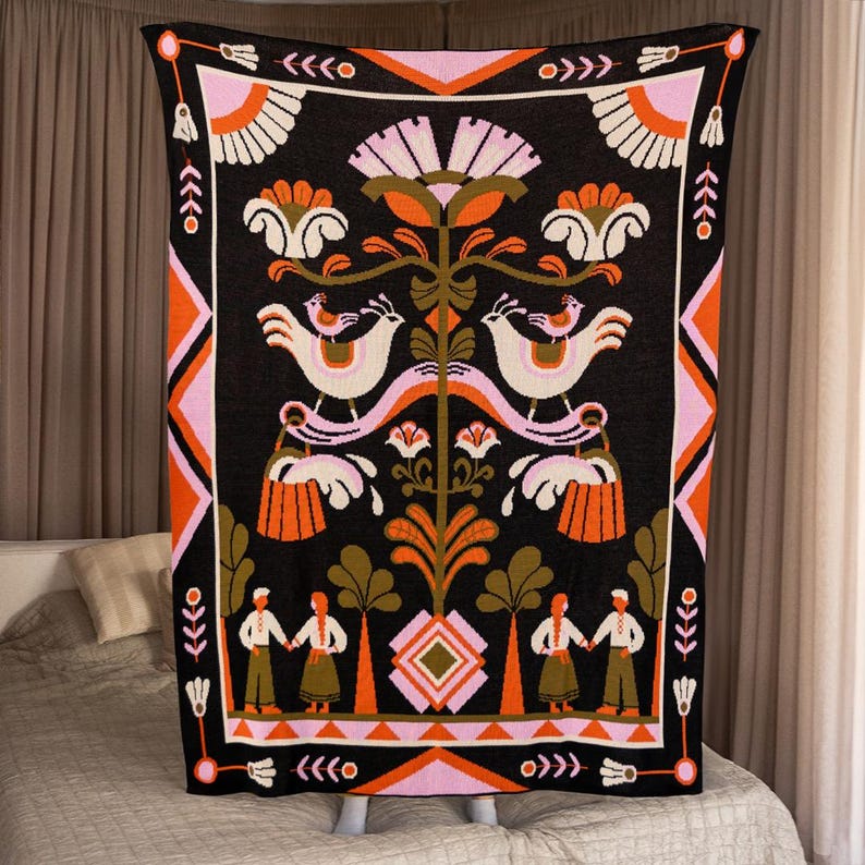 May include: A black blanket with a colorful folk art design. The blanket features white birds, orange and pink flowers, and figures holding hands. The border has geometric patterns in orange, pink, and black. The blanket is displayed on a bed.