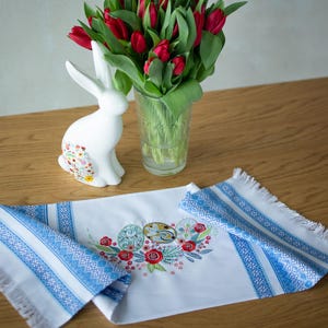 Peut inclure: Une figurine de lapin en céramique blanche avec un motif floral, un vase de tulipes rouges et un chemin de table blanc avec des rayures bleues et des œufs de Pâques brodés.