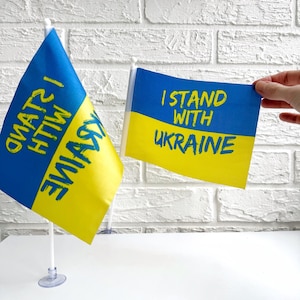 Può includere: Due piccole bandiere con i colori della bandiera ucraina. Una bandiera dice "Sto con l'Ucraina" e l'altra dice "Sto con l'Ucraina" in ucraino.