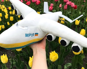 Puede incluir: Un avión de peluche blanco con la palabra "MPIR" y la bandera de Ucrania impresa en el lateral. El juguete se sostiene en una mano sobre un fondo de tulipanes rosas y amarillos.