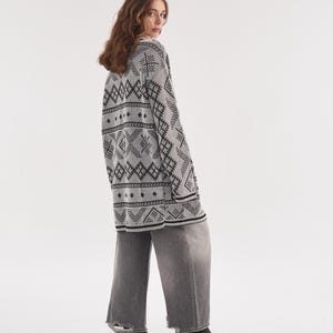 May include: An oversized white cardigan featuring a black geometric pattern, long sleeves, and a V-neckline, paired with wide-leg gray denim jeans that have frayed hems and black pointed-toe ankle boots with a small heel.