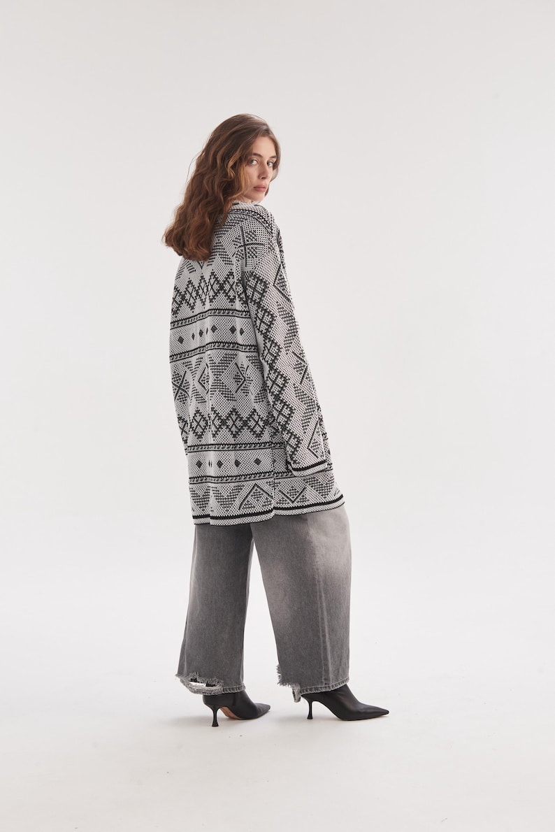May include: An oversized white cardigan featuring a black geometric pattern, long sleeves, and a V-neckline, paired with wide-leg gray denim jeans that have frayed hems and black pointed-toe ankle boots with a small heel.