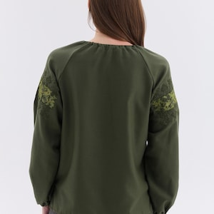 May include: Olive green long-sleeve blouse with embroidered floral design on the sleeves. The blouse has a round neckline and a relaxed fit. The sleeves are long and gathered at the wrists. The back of the blouse is visible.