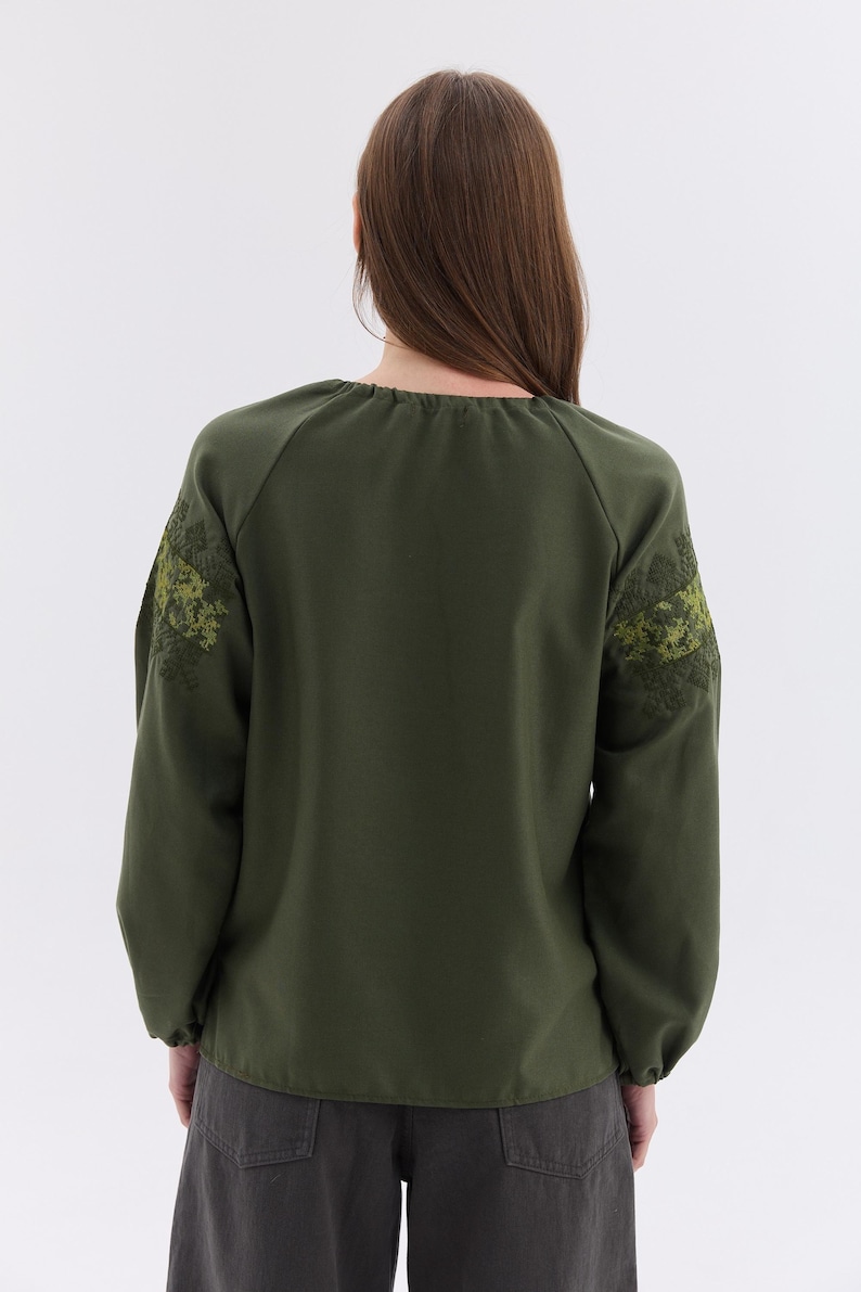 May include: Olive green long-sleeve blouse with embroidered floral design on the sleeves. The blouse has a round neckline and a relaxed fit. The sleeves are long and gathered at the wrists. The back of the blouse is visible.