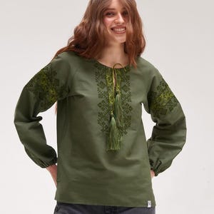 May include: Olive green long-sleeve blouse with embroidered floral patterns on the sleeves and chest. The blouse features a V-neck with a tassel tie and gathered cuffs. The sleeves are long and loose, and the fabric appears to be linen or a similar material.