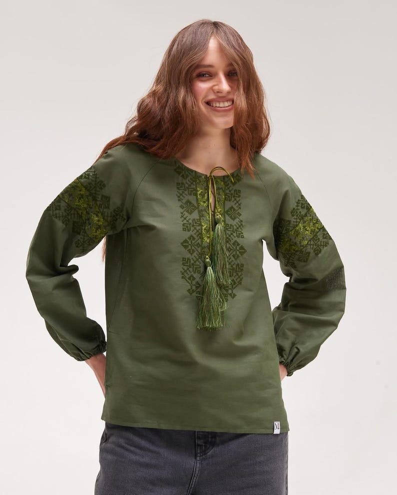 May include: Olive green long-sleeve blouse with embroidered floral patterns on the sleeves and chest. The blouse features a V-neck with a tassel tie and gathered cuffs. The sleeves are long and loose, and the fabric appears to be linen or a similar material.