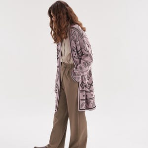 May include: A long-sleeved, open-front cardigan in a light pink hue featuring a dark brown geometric pattern. The cardigan is paired with a light beige button-down shirt, loose-fitting olive-toned trousers, and dark brown slip-on shoes.