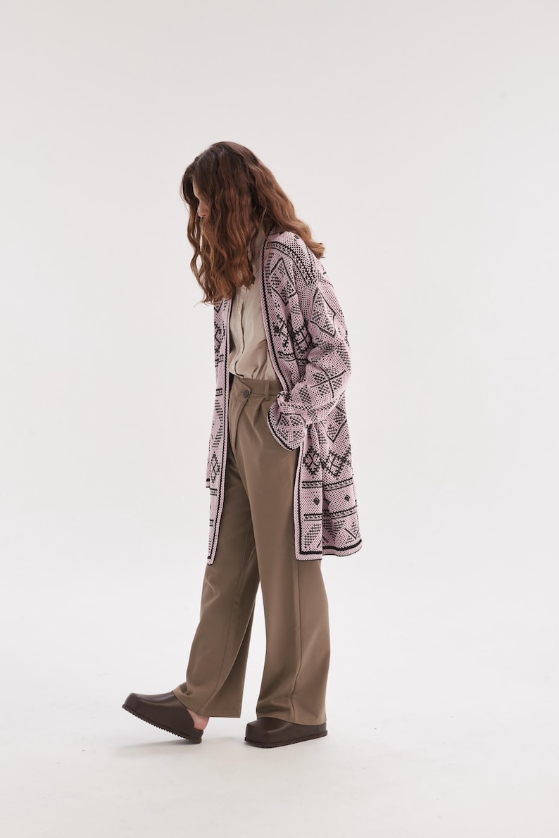May include: A long-sleeved, open-front cardigan in a light pink hue featuring a dark brown geometric pattern. The cardigan is paired with a light beige button-down shirt, loose-fitting olive-toned trousers, and dark brown slip-on shoes.