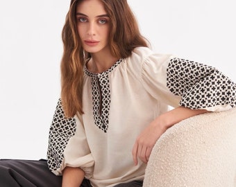 May include: A white cotton blouse with black embroidered details on the sleeves and neckline. The blouse has a relaxed fit and a slightly gathered neckline.
