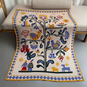 May include: A white throw blanket with a colorful folk art design featuring a tree with birds, flowers, and a rooster. The blanket has a blue and yellow border.