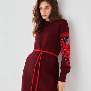May include: A burgundy sweater dress with long sleeves and a red embroidered floral pattern on the sleeves. The dress has a red tassel tie belt.