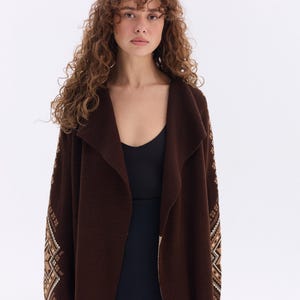 May include: A long, dark brown cardigan with a shawl collar and patterned sleeves. The sleeves feature a geometric design in shades of beige and brown. The cardigan is worn over a black top.