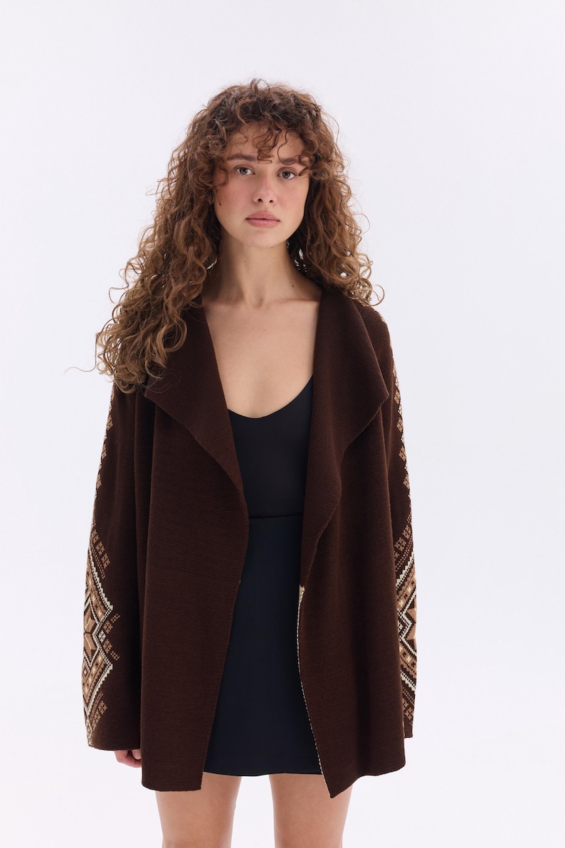 May include: A long, dark brown cardigan with a shawl collar and patterned sleeves. The sleeves feature a geometric design in shades of beige and brown. The cardigan is worn over a black top.