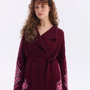May include: A burgundy wrap cardigan with a shawl collar and a matching belt. The sleeves feature a pink and white embroidered design. The cardigan is paired with white pants.