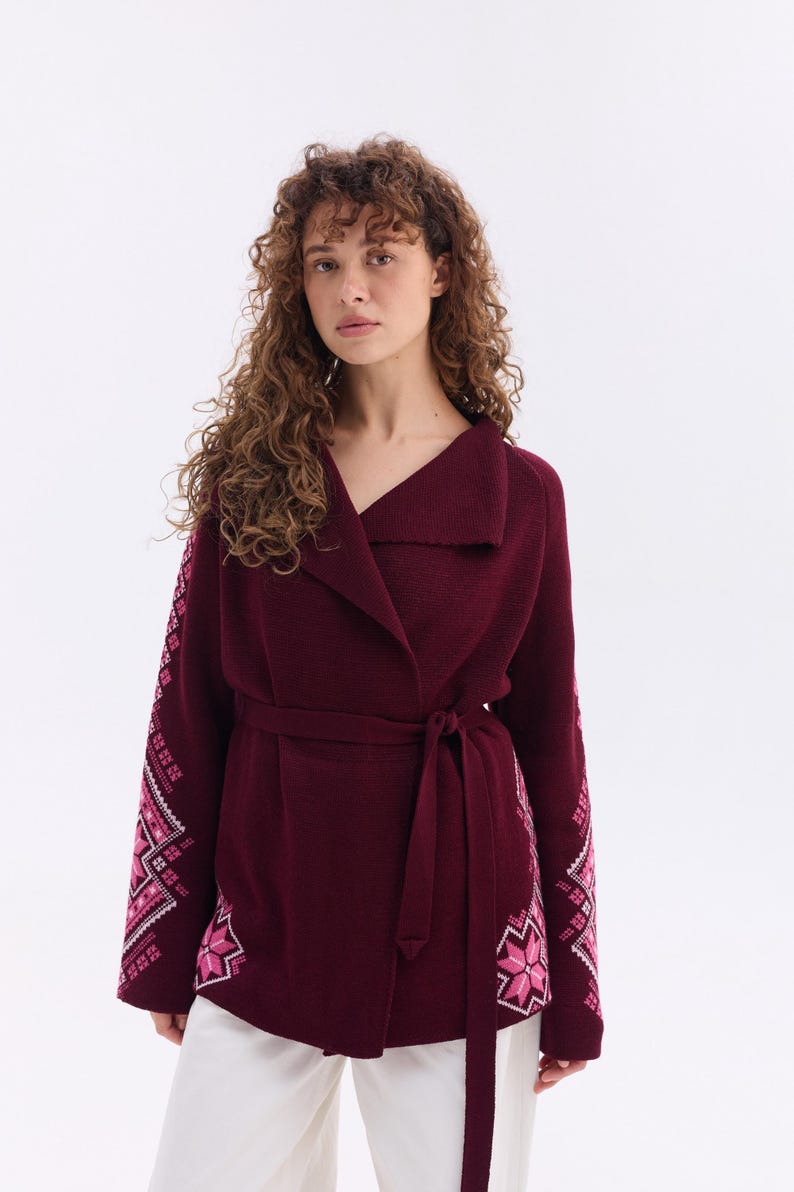 May include: A burgundy wrap cardigan with a shawl collar and a matching belt. The sleeves feature a pink and white embroidered design. The cardigan is paired with white pants.