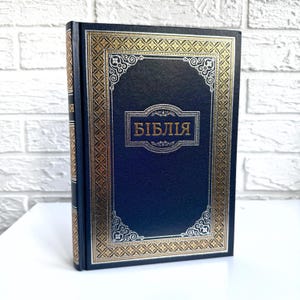 May include: A navy blue hardback book with a gold and silver decorative border. The title "Библия" is printed in gold on the front cover.