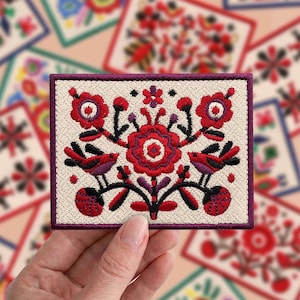 Könnte beinhalten: Ein rechteckiger, bestickter Aufnäher mit cremefarbenem Hintergrund und einem symmetrischen Volkskunstmotiv. Das Design zeigt eine zentrale rote Blume, zwei einander zugewandte Vögel sowie florale Elemente in Rot, Schwarz und Violett.