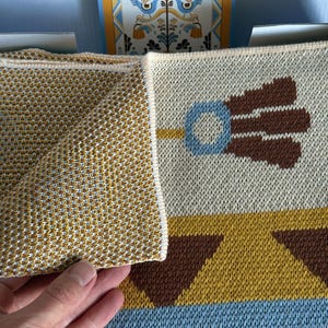 May include: A folded, patterned blanket with a geometric design in shades of gold, brown, blue, and cream. The blanket features a repeating pattern of stylized floral and geometric shapes. The edge of the blanket is a solid gold color.