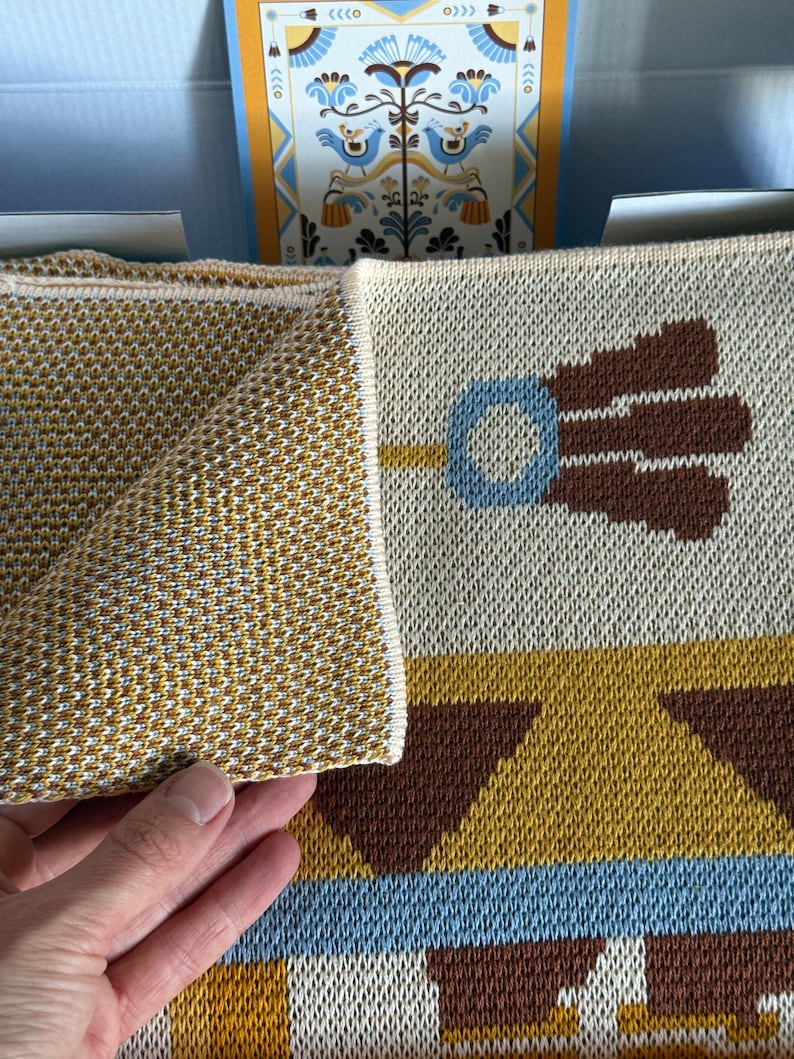 May include: A folded, patterned blanket with a geometric design in shades of gold, brown, blue, and cream. The blanket features a repeating pattern of stylized floral and geometric shapes. The edge of the blanket is a solid gold color.