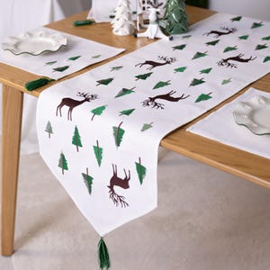 May include: White table runner with a repeating pattern of brown deer and green Christmas trees. The runner has green tassels on each end. Matching placemats and plates are also visible on the wooden table.