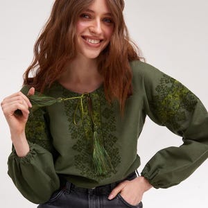 May include: Olive green embroidered blouse with long sleeves and a drawstring neckline. The blouse features intricate green embroidery on the sleeves and bodice. The sleeves are gathered at the cuffs. The model is wearing dark gray jeans.