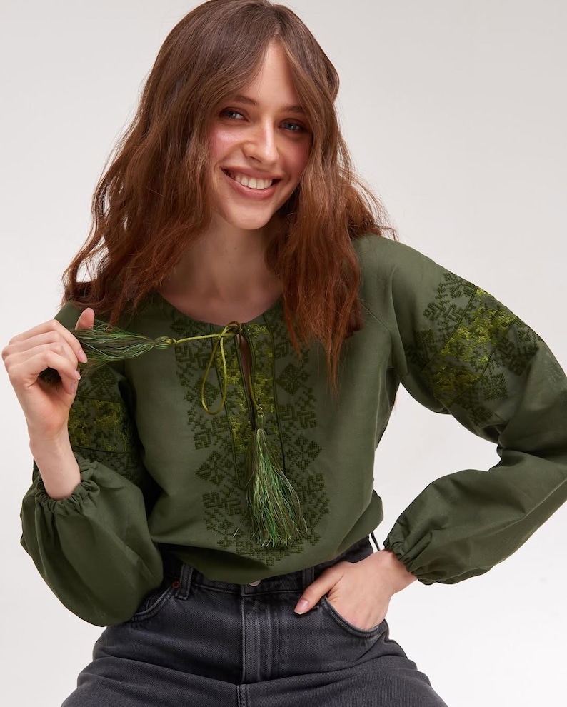 May include: Olive green embroidered blouse with long sleeves and a drawstring neckline. The blouse features intricate green embroidery on the sleeves and bodice. The sleeves are gathered at the cuffs. The model is wearing dark gray jeans.