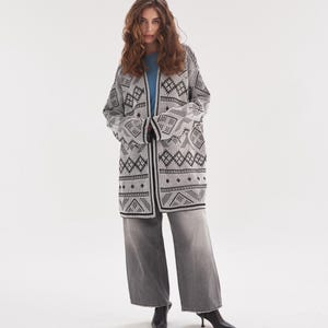 May include: A long-sleeved, open-front cardigan featuring a black and white geometric pattern, worn over a solid light blue t-shirt. The outfit includes wide-leg, faded grey denim jeans and black pointed-toe ankle boots with a small heel.