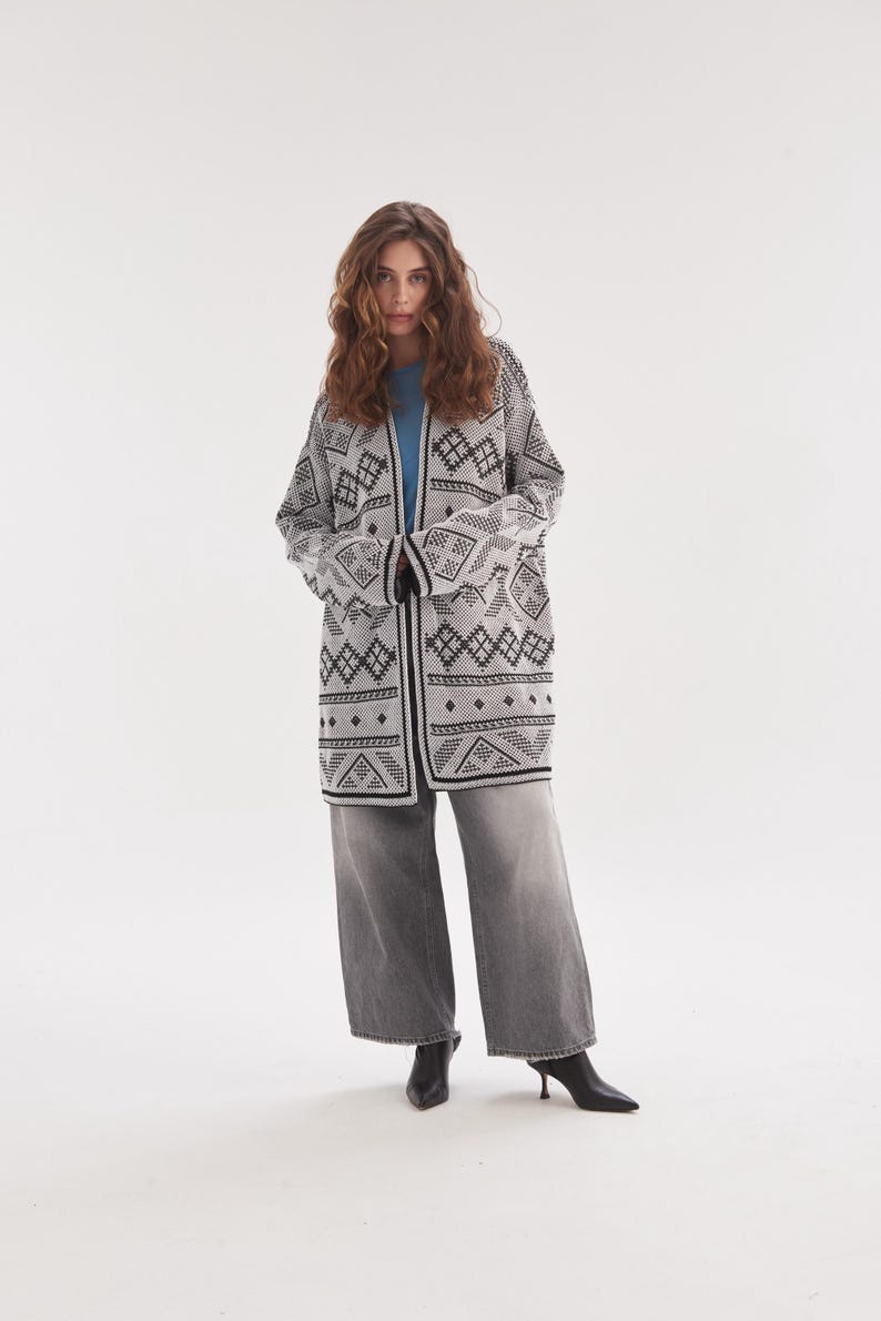 May include: A long-sleeved, open-front cardigan featuring a black and white geometric pattern, worn over a solid light blue t-shirt. The outfit includes wide-leg, faded grey denim jeans and black pointed-toe ankle boots with a small heel.