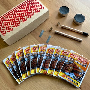 Peut inclure: Un kit d'artisanat avec une boîte en carton ornée d'un motif géométrique rouge et blanc. Le kit comprend des sachets avec du texte cyrillique, deux petits récipients, deux outils avec des manches en bois et des pièces métalliques.