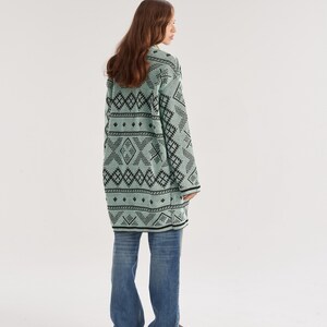 May include: A long-sleeved, mint green cardigan featuring a repeating black geometric pattern of diamonds and horizontal lines, paired with loose-fitting blue denim jeans and dark brown slip-on shoes, viewed from the back.