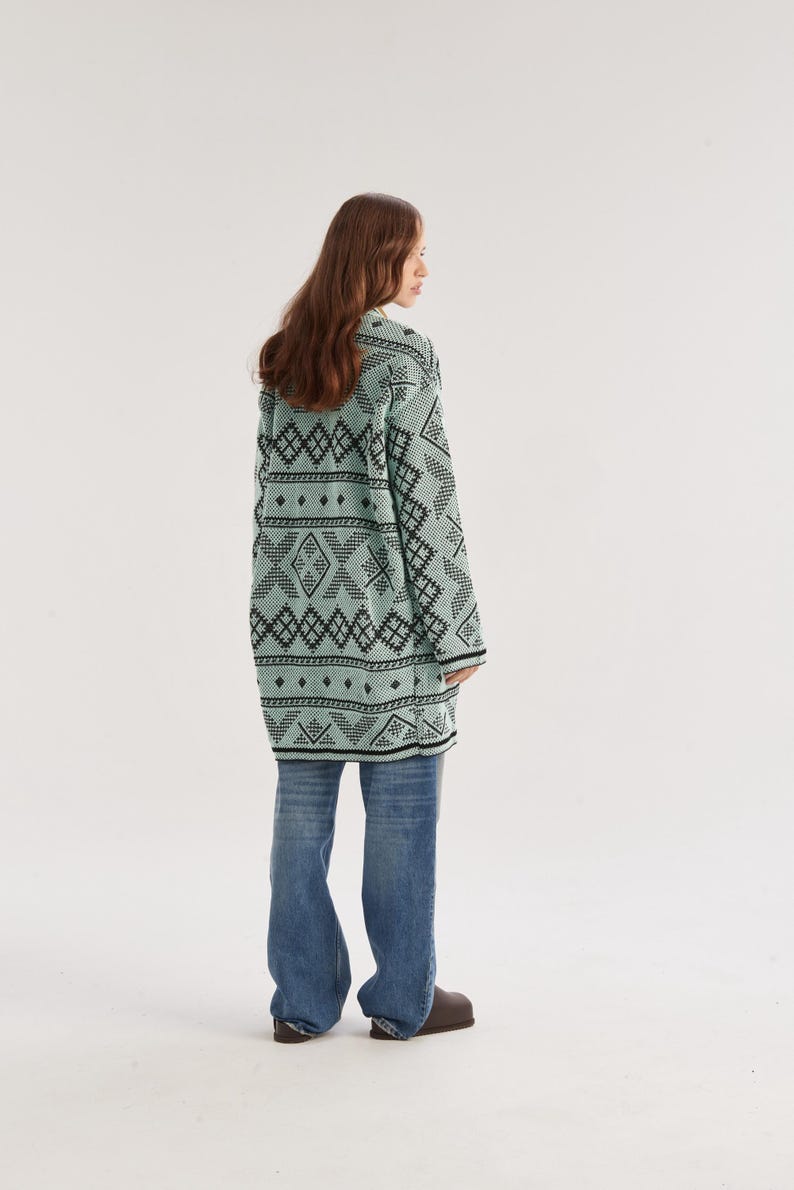 May include: A long-sleeved, mint green cardigan featuring a repeating black geometric pattern of diamonds and horizontal lines, paired with loose-fitting blue denim jeans and dark brown slip-on shoes, viewed from the back.
