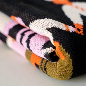 May include: Folded knit fabric with a repeating pattern of white, black, pink, and orange shapes. The fabric has a textured appearance, with visible knit stitches. The colors are vibrant and the design is eye-catching.