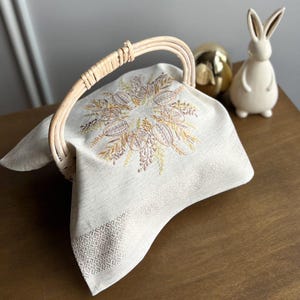 May include: A wicker basket with a curved handle contains a cream linen cloth. The cloth features an embroidered floral design in gold and brown. A ceramic rabbit and a gold egg are in the background.