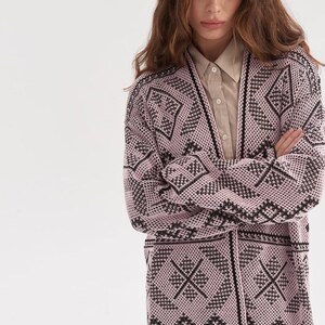 May include: A long-sleeved, open-front cardigan featuring a light pink base with a repeating black geometric pattern. The knit garment has a V-neckline and is worn over a beige button-down shirt with a pointed collar and visible buttons.