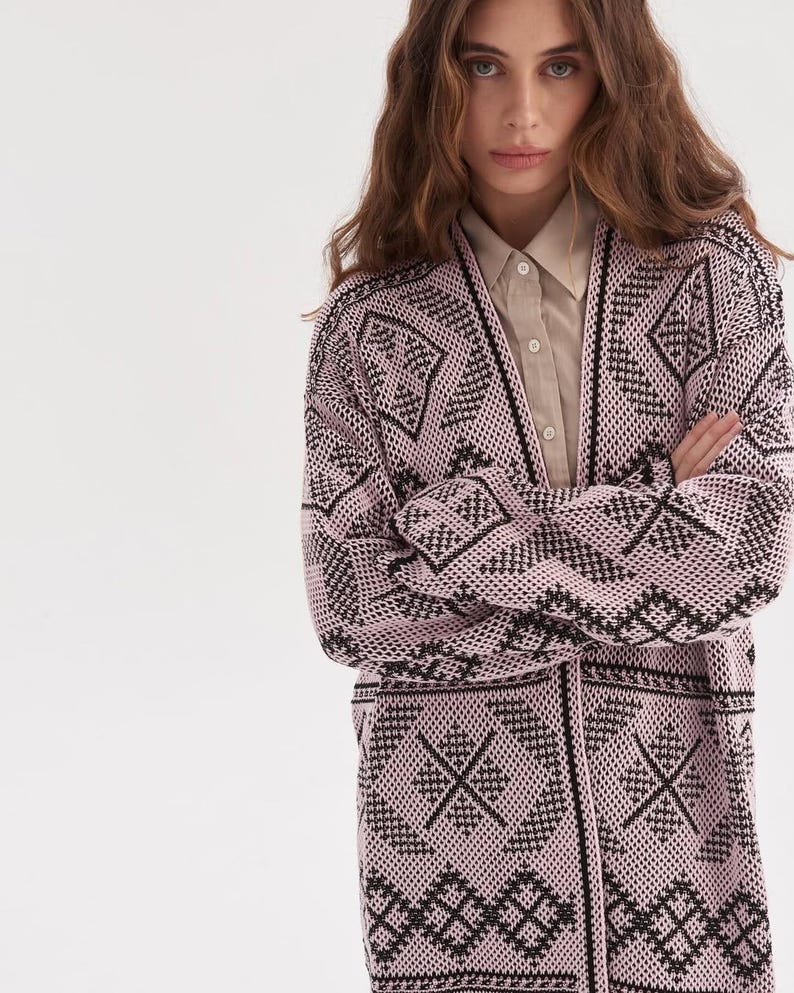 May include: A long-sleeved, open-front cardigan featuring a light pink base with a repeating black geometric pattern. The knit garment has a V-neckline and is worn over a beige button-down shirt with a pointed collar and visible buttons.