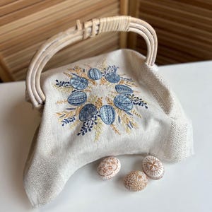 以下が含まれることがあります： 白のリネン製のイースターバスケットで、織り込まれたハンドルと青と黄色の刺繍された花柄が施されています。バスケットには、装飾されたイースターエッグが3つ入っています。