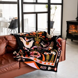May include: A black throw blanket with a colorful folk art design featuring birds, trees, and figures. The blanket is draped over a brown leather sofa. The design includes pink, orange, white, and green accents.