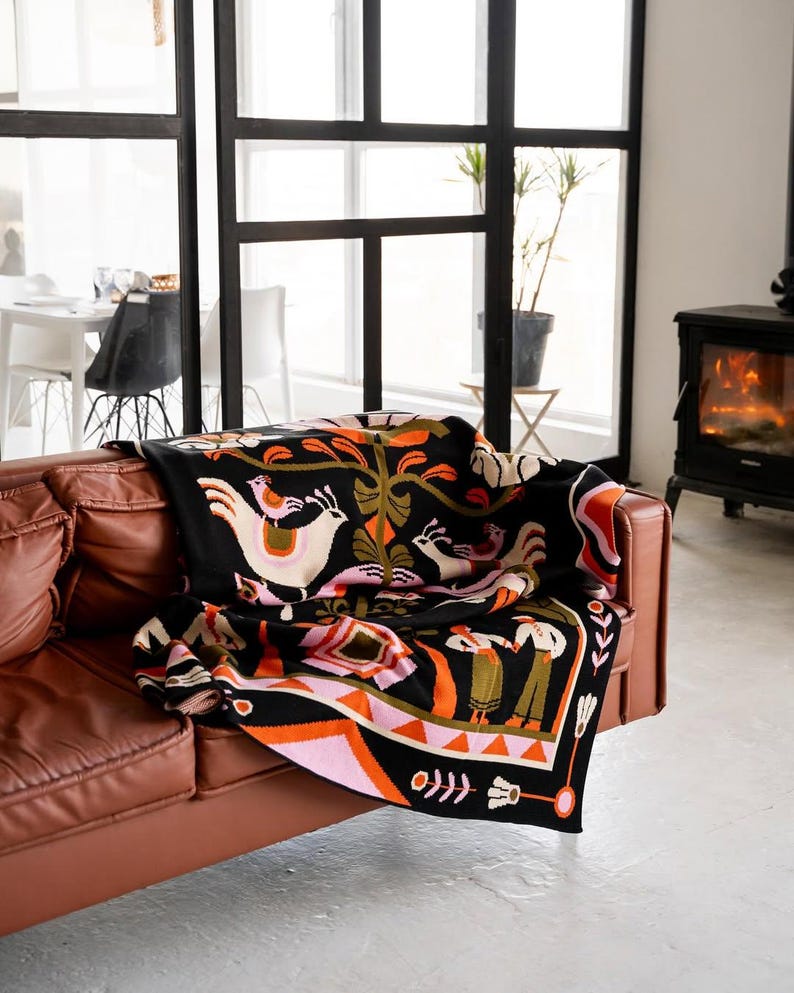 May include: A black throw blanket with a colorful folk art design featuring birds, trees, and figures. The blanket is draped over a brown leather sofa. The design includes pink, orange, white, and green accents.