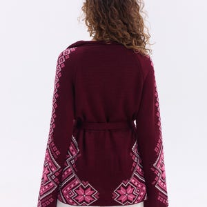 May include: Burgundy cardigan with a matching belt and intricate pink and white geometric patterns on the sleeves and lower front. The sweater has a shawl collar and is worn with white pants. A stylish, warm garment.