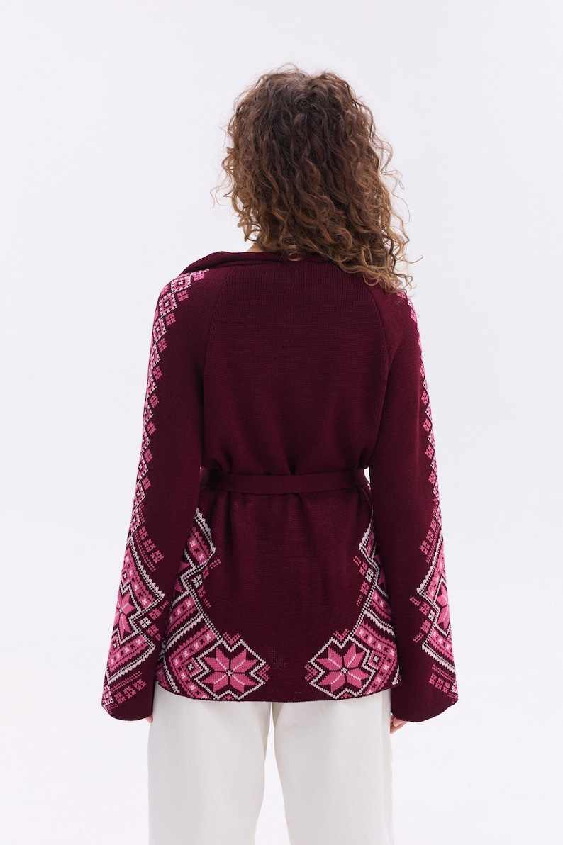 May include: Burgundy cardigan with a matching belt and intricate pink and white geometric patterns on the sleeves and lower front. The sweater has a shawl collar and is worn with white pants. A stylish, warm garment.