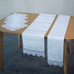 May include: White lace table runners and matching square doilies are displayed on a wooden table. The runners have a detailed lace trim and a patterned design. The doilies are also white with a scalloped edge.