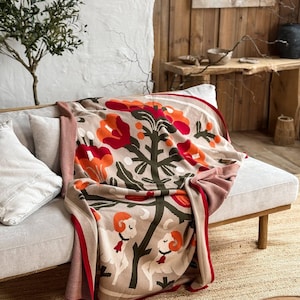 May include: A beige throw blanket with a red border and a pattern of orange flowers, green leaves, and white sheep. The blanket is draped over a light gray sofa with white pillows. The throw is made of a soft material.