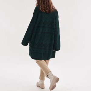 May include: An oversized dark green knit cardigan featuring a black geometric diamond pattern, paired with beige trousers and light-colored lace-up boots. The long-sleeved garment has a relaxed fit and reaches mid-thigh, showcasing a textured knit.