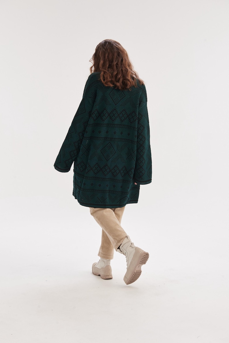 May include: An oversized dark green knit cardigan featuring a black geometric diamond pattern, paired with beige trousers and light-colored lace-up boots. The long-sleeved garment has a relaxed fit and reaches mid-thigh, showcasing a textured knit.