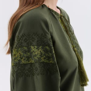 May include: Olive green tunic with embroidered details on the sleeves. The tunic features a round neckline with a drawstring and a long tassel. The sleeves have a geometric pattern in shades of green.
