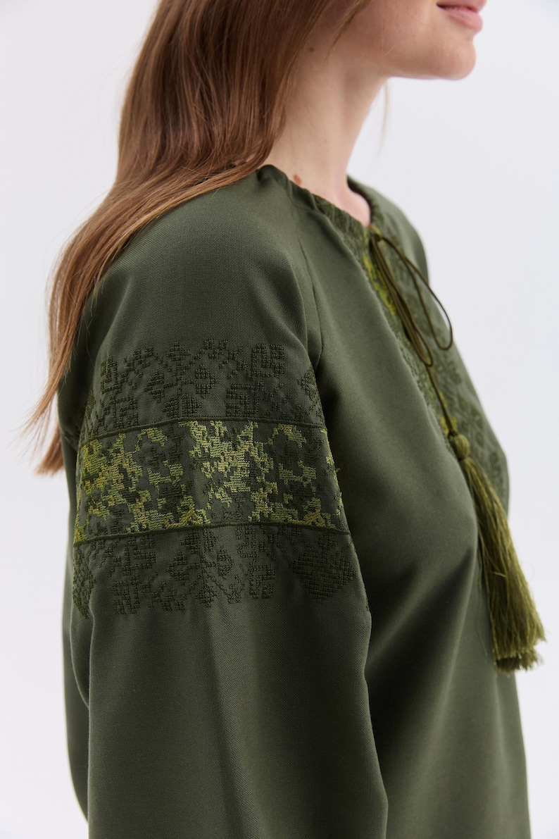 May include: Olive green tunic with embroidered details on the sleeves. The tunic features a round neckline with a drawstring and a long tassel. The sleeves have a geometric pattern in shades of green.