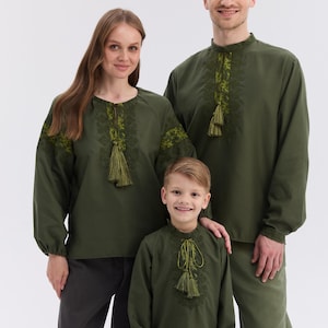 May include: Three people wearing olive green embroidered shirts with long sleeves and tassel details. The shirts have intricate floral embroidery around the neckline and sleeves. The family is standing in front of a white background.