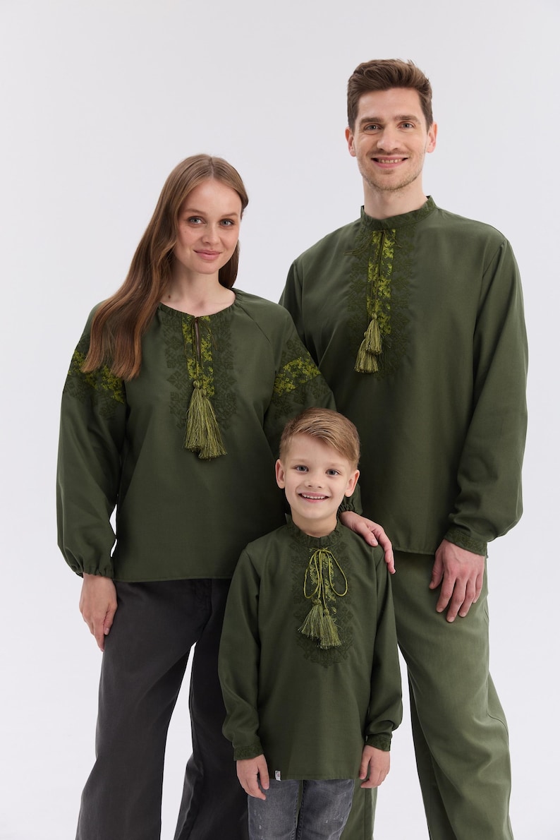 May include: Three people wearing olive green embroidered shirts with long sleeves and tassel details. The shirts have intricate floral embroidery around the neckline and sleeves. The family is standing in front of a white background.