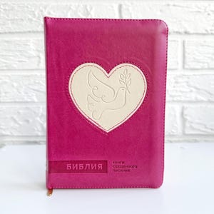 Könnte beinhalten: Eine rosafarbene, ledergebundene Bibel mit einem weißen, geprägten Herzen, das eine Taube und einen Olivenzweig zeigt. Der Text "Библия" und "Книги Священного Писания" ist auf dem Einband aufgedruckt.